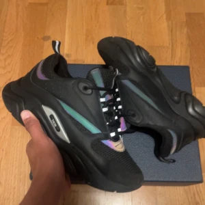 Svarta B22 sneakers med iriserande detaljer - Säljer ett par chunky  B22 sneakers i svart med coola iriserande detaljer i grönt, lila och blått. Skorna har mesh och skinnpaneler, svart snörning och en rejäl platt sula. Perfekta för dig som vill sticka ut med en lyxig och modern look.