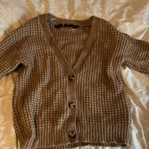 Beige stickad kofta med knappar - Mysig beige stickad kofta från Lager 157 med grovstickat mönster och tre stora knappar framtill. Djup v-ringning och ribbade muddar ger en chill vibe. Perfekt att slänga över en t-shirt när det är kyligt ute.