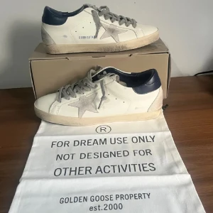 Golden Goose vita sneakers med grå stjärna - Golden Goose sneakers i vitt skinn med grå mockadetaljer och en stor grå stjärna på sidan. Skorna har grå snörning, mörkblå häl och platt sula. Snyggt slitna detaljer och klassisk rund tå. Perfekt för dig som gillar unik design och streetstyle.