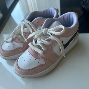 Nike Air Jordan 1 Low rosa/vit - Säljer ett par Nike Air Jordan 1 Low sneakers i en snygg kombination av ljusrosa och vitt med svart swoosh. Skorna har klassisk snörning, perforerad tå och platt sula. Materialet är skinn och textil, och de har rund tå. Perfekt för dig som gillar streetstyle.