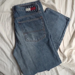 Vintage Tommy Jeans raka 32/32 - Klassiska blå jeans från Tommy Jeans med raka ben och normal passform. Snyggt broderad logga bak och detaljer i rött, vitt och blått vid linningen. Tillverkade i slitstarkt bomullsjeansmaterial. Perfekta för en avslappnad och stilren look.