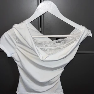 Vit topp med spets från SHEIN - Säljer en vit topp från SHEIN i storlek XS. Toppen har korta ärmar och en snygg draperad halsringning med transparent spetsdetalj framtill. Materialet är mjukt och stretchigt, perfekt för dig som gillar en feminin och trendig look.