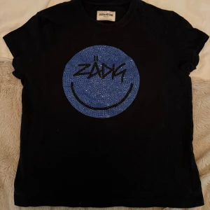 Svart Zadig & Voltaire t-shirt med blått tryck - Cool svart t-shirt från Zadig & Voltaire med ett stort blått smiley-tryck och glittriga detaljer på bröstet. T-shirten har korta ärmar och rund hals. Perfekt för dig som gillar statement-plagg och vill sticka ut med en unik design. Mycket bra skick och storlek S