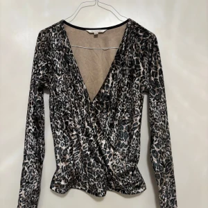 Leopardmönstrad omlottopp från First Class Party - Säljer en snygg leopardmönstrad omlottopp från First Class Party i storlek S. Toppen har lång ärm, v-ringad omlottfront och är gjord i glansigt syntetmaterial som ger en cool look. Perfekt för dig som vill sticka ut med djurmönster och trendig stil.