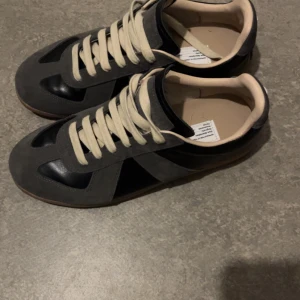 Grå och svarta sneakers Maison Margiela - Snygga sneakers från Maison Margiela i grå mocka och svart skinn med beige snörning. Har använt dem sparsamt och dem är storlek 41