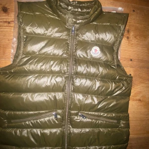 Olivgrön dunväst från Moncler - Säljer en olivgrön dunväst från Moncler med hög krage och två dragkedjefickor framtill. Västen har quiltad design och klassisk Moncler-logga på bröstet. Perfekt att slänga över en hoodie eller tröja för extra värme. Storlek M/2 prislapp och påse fås med. Tveka ej att fråga eller buda!