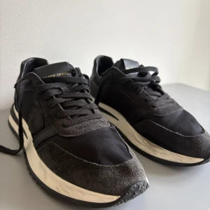 Svarta sneakers från Philippe Model - Snygga svarta sneakers från Philippe Model med detaljer i mocka och nylon. Skorna har rund tå, snörning och vit platt sula med svarta inslag. Stilren design med diskret logga på plösen och insidan. Perfekta för dig som gillar klassisk streetstil. Storleken är 39 men är mer som 40 eller 40,5.                             Prisk kan diskuteras!