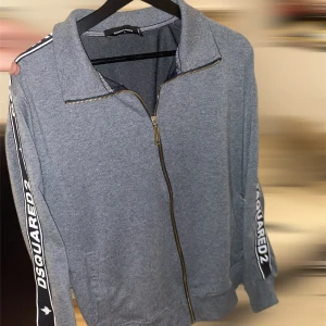 Grå ziptröja från Dsquared2 - Snygg grå ziptröja från Dsquared2 med dragkedja framtill och svarta ränder med vit logga längs ärmarna. Storlek L men passar M skulle jag säga. Kom gärna privat för mer frågor!
