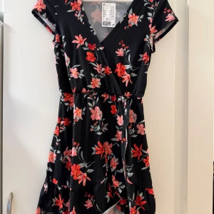 Svart blommig kortklänning H&M - Supersöt svart kortklänning från H&M med färgglada blommor i rött och rosa. Klänningen har omlott-v-ringning, korta ärmar och volangdetalj nertill. Tillverkad i mjuk polyester med lite stretch för extra komfort. Perfekt för dig som gillar en feminin och trendig stil.