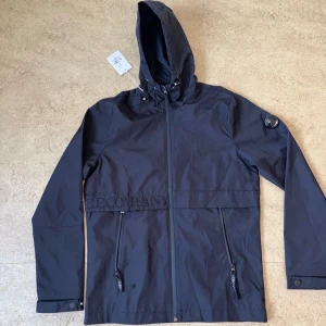  C.P. Company vindkacka  - Svart vindjacka från C.P. Company med huva, dragkedja framtill och två snedställda fickor med dragkedja. Jackan har den klassiska linsen på ärmen och broderad logga över bröstet. Perfekt för dig som gillar stilren och sportig look.