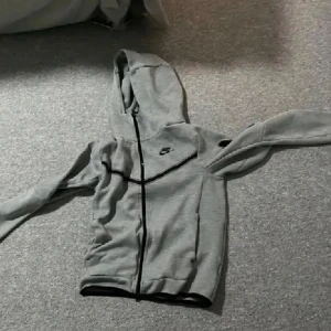 Grå hoodie från Nike med dragkedja - Säljer en grå hoodie från Nike med hel dragkedja och huva. Tröjan har långa ärmar, svarta detaljer och en klassisk Nike-logga på bröstet. Perfekt för chill dagar eller när du vill ha en sportig look. 