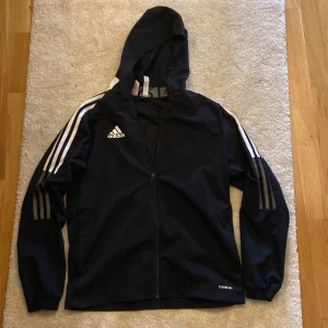 Svart Adidas vindjacka med huva - Svart vindjacka/tunn kofta från Adidas med klassiska vita ränder längs ärmarna och huva. Jackan har hel dragkedja framtill och Aeroready-tryck vid nederkanten. Materialet är lätt och syntetiskt, perfekt för sport och träning. Snyggt Adidas-logga på bröstet och långärmad modell.