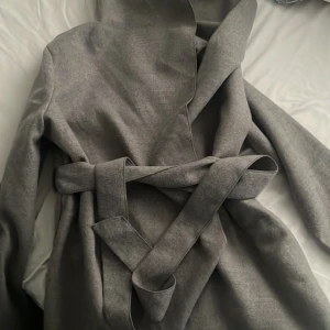 Grå omlottkappa med huva och bälte - Snygg grå kappa med omlottmodell och breda slag, perfekt för dig som gillar stilren look. Jackan har en stor huva och ett matchande bälte i midjan för att skapa en cool siluett. Materialet är mjukt och känns skönt mot huden. Passar dig som vill ha en chill och trendig vibe.