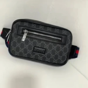 Svart axelväska från Gucci med mönster - Snygg svart axelväska från Gucci med klassiskt GG-mönster och silverfärgade dragkedjor. Väskan har en rektangulär form och bred axelrem i blått och rött. Materialet är en mix av skinn och syntet, perfekt för att lyfta din outfit.