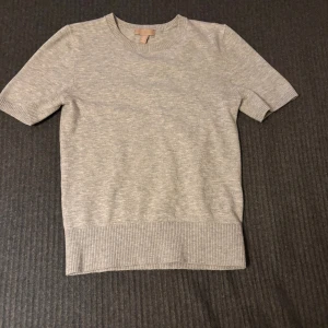 Grå stickad kortärmad tröja H&M XS - En stilren ljusgrå stickad tröja från H&M i storlek XS. Tröjan har korta ärmar, rund halsringning och bred ribbad kant nedtill och vid ärmsluten. Perfekt för lager-på-lager och enkel att matcha med olika outfits. På bilden syns en smink fläck men alla mina plagg tvättas innan de postas och fläcken kommer försvinna i tvätten, även en söm som har lossnat men det syns självklart inte på. 