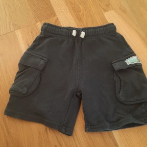 Svarta shorts med fickor från Zara - Svarta shorts från Zara med elastisk midja och snörning. Shortsen har två stora cargofickor på sidorna och en liten patchdetalj på ena benet. Tillverkade i mjuk bomull, perfekta för varma dagar.