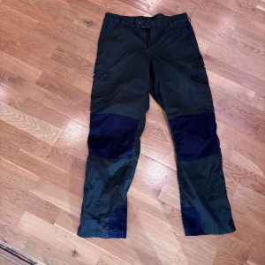 Mörkgröna cargopants från Pinewood - Säljer ett par mörkgröna cargopants från Pinewood Outdoor Collection. Byxorna har svarta förstärkningar på knän, bak och benslut, samt flera praktiska fickor med lock och knapp. Perfekta för uteliv och äventyr. Resår i midjan för extra komfort.