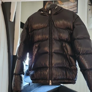 Svart pufferjacka från Zara S - Svart pufferjacka från Zara i storlek S. Jackan har en glansig finish, huva med snörning och dragkedja framtill. Den är vadderad och har en loose passform som ger en cool och avslappnad vibe. Perfekt för kalla dagar när du vill vara både varm och snygg.