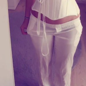 Vita satinbyxor med snörning - Snygga vita byxor i satin med låg midja och avslappnad passform. Byxorna har snörning i midjan och fickor på sidorna. Materialet är glansigt och mjukt, vilket ger en cool och trendig vibe. Perfekta för dig som gillar att sticka ut med din stil.