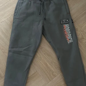 Grå Hoodrich mjukisbyxor med tryck - Snygga grå mjukisbyxor från Hoodrich med coolt tryck och logga i vitt och rött på benen. Byxorna har snörning i midjan, ribbade muddar och sidofickor. Perfekta för chill eller streetwear-stil. Materialet är mjukt och bekvämt.