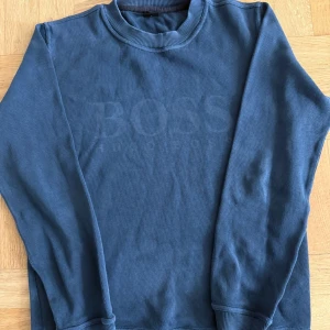 Blå sweatshirt från Hugo Boss - Säljer en stilren blå sweatshirt från Hugo Boss med diskret logga framtill. Tröjan har rund hals, ribbade muddar och är tillverkad i mjuk bomull. Perfekt för en chill och snygg look.