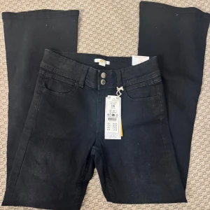 Svarta bootcut jeans med hög midja - Svarta jeans med bootcut-modell och hög midja. Byxorna har klassisk femficksdesign, dubbla knappar i midjan och raka ben med lätt utsvängning. Materialet är jeans och färgen är helsvart, perfekt för en stilren look.