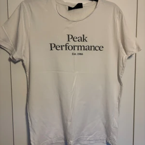 Vit Peak Performance t-shirt i bomull - Vit t-shirt från Peak Performance med svart tryck framtill. Klassisk rund halsringning och korta ärmar. Tillverkad i mjuk bomull för en skön känsla. Perfekt för en clean och stilren look.
