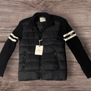 Svart Moncler pufferjacka med stickade ärmar - Svart pufferjacka från Moncler med stickade ärmar och vita ränder. Jackan har dragkedja framtill, två dragkedjefickor och Moncler-logga på ena ärmen. Kroppen är quiltad och glansig medan ärmarna är kabelstickade för en snygg kontrast.
