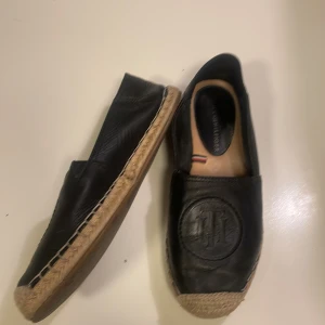 Svarta espadrillos från Tommy Hilfiger - Snygga svarta espadrillos från Tommy Hilfiger i skinn med tydlig logga framtill. Skorna har klassisk flätad sula i naturfärgad jute och små Hilfiger-detaljer på insidan och hälen. Perfekta för en chill sommarlook.