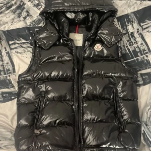  Moncler väst   - Säljer en svart, glansig dunväst från Moncler med huva och dragkedja framtill. Västen har två fickor med dragkedja och klassisk Moncler-logga på bröstet. Perfekt för lager-på-lager och snygg till streetwear. Fodrad och quiltad för extra värme.