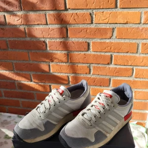 Adidas sneakers i grått och vitt EUR 36 2/3 - Säljer ett par klassiska Adidas sneakers med retrovibbar i grått, vitt och röda detaljer. Skorna har de ikoniska tre ränderna på sidan, snörning och platt sula. Ovandelen är i textil och mocka, med Adidas-logga på häl och plös. Perfekt för dig som gillar old school-stil.