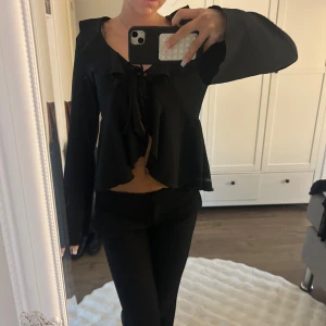 Svart blus med volang och snörning - Säljer en svart blus med långa ärmar och snygg volangkant längs halsringningen. Blusen har snörning framtill som ger en cool och feminin touch. Materialet känns lätt och flowy, perfekt för dig som gillar en lite romantisk vibe.