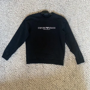 Svart sweatshirt Emporio Armani M - Svart sweatshirt från Emporio Armani med vit logga och text på bröstet. Klassisk rund halsringning och långa ärmar. Tröjan är i mjuk bomull och har en stilren, enkel design som passar perfekt till jeans eller joggers. Litet hål på baksidan. 