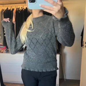 Grå stickad tröja med detaljer  - Mysig grå stickad tröja med diskret rutmönster och ribbade muddar. Tröjan har lång ärm och en klassisk rund halsringning. Perfekt för lager-på-lager och chill dagar.