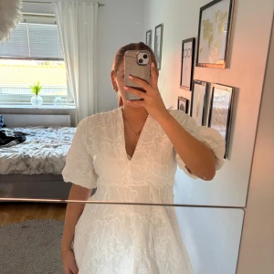 Studentklänning  - Superfin vit kort klänning med v-ringning och puffiga korta ärmar. Klänningen har ett broderat blommigt mönster över hela tyget som ger en lyxig känsla. Passformen är normal och tyget känns lätt och luftigt, perfekt för varma dagar. Snygg detalj med volym i ärmarna.