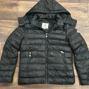 Svart dunjacka moncler - Säljer en svart dunjacka från Moncler i storlek S. Jackan har quiltad design, avtagbar huva och två snedställda fickor med dragkedja. Klassisk Moncler-logga på ärmen och blanka detaljer. Perfekt för kalla dagar och riktigt snygg streetstil.