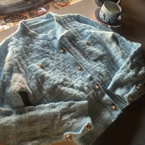 Ljusblå kofta med pärlknappar - Superfin ljusblå kofta med glittrig struktur och dekorativa pärlknappar. Koftan har krage, ribbade muddar och ett rutigt mönster i stickad kvalitet. Perfekt för dig som gillar en chic och mysig look.