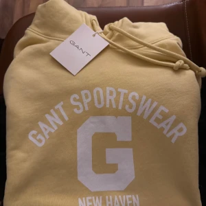 Gul hoodie från Gant med tryck - Gul hoodie från Gant med vit tryckt text 'GANT SPORTSWEAR NEW HAVEN 1949' och ett stort G på bröstet. Tröjan har huva med snörning och ribbade muddar. Perfekt för en chill och sportig look. Tröjan är helt ny och jag säljer den på grund av att jag har ångrat mig. 