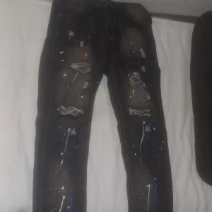 Svarta jeans med färgstänk och slitningar - Säljer ett par svarta jeans med coola färgstänk i vitt, blått och rött samt slitna detaljer och hål på benen. Jeansen har en smal passform och är perfekta för dig som vill sticka ut med en edgy streetstyle.