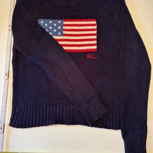 Mörkblå stickad tröja Polo Ralph Lauren - Mörkblå stickad tröja från Polo Ralph Lauren med amerikansk flagga framtill och RL-broderi. Tröjan har rund hals, ribbade muddar och lång ärm. Perfekt för dig som gillar klassisk preppy stil och vill sticka ut med en ikonisk detalj.