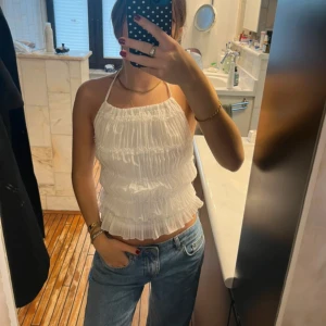 Vit plisserad linnetopp med halterneck - Säljer en vit linnetopp med halterneck och plisserade volanger. Toppen är ärmlös och har en lätt croppad passform som ger en fräsch och somrig vibe. Perfekt för varma dagar och enkel att styla med jeans eller kjol.