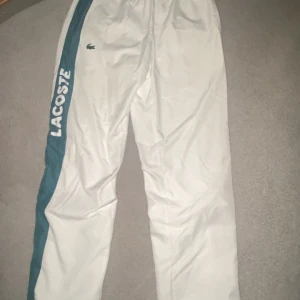 Vita Lacoste Sport byxor XS - Säljer ett par vita Lacoste Sport byxor i storlek XS. Byxorna har en blågrön och en röd sidopanel, samt stor Lacoste-logga på benet och klassisk krokodil på framsidan. De har elastisk midja och är gjorda i ett lätt, syntetiskt material med sportig känsla.