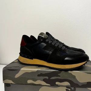 Valentino Garavani Rockrunner  - Snygga Valentino Garavani Rockrunner sneakers i svart och mörkblått med camouflagemönster, röda detaljer på hälen. Ny skick box ingår storlek 42 en blandning av svart o mörkblå perfekt nu till dem kallare tiderna