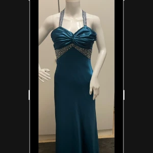 Bal/fest klänning - Fin elegant fest/bal klänning med "diamant pärlor", banden går som ett kors och har ganska öppen ryggen, men den är super snygg! Har bara använt den 1 gång på min bal så den är väldigt bra bevarad. Färgen är en djup petrolblå eller mörk teal-satin färg — en blandning mellan mörkblå och grön med en metallisk, nästan silkeslen glans. Under vissa ljus (särskilt varmvitt ljus) kan den dra mer åt mörk turkos eller smaragdblå, medan kallare ljus får den att se mer djupblå ut.