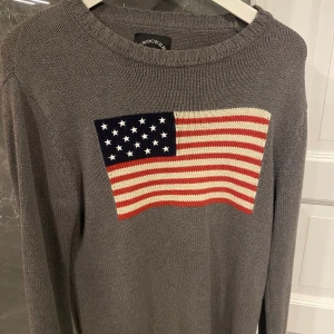 Grå stickad tröja Crocker - Snygg grå stickad tröja från Crocker i storlek XS. På bröstet finns ett stort amerikanskt flaggmotiv i blått, vitt och rött. Tröjan har rund hals och långa ärmar, perfekt för dig som gillar klassisk stil med en twist.