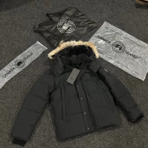 Svart Canada Goose jacka med päls - Svart äkta jacka från Canada Goose med fluffig päls på huvan och klassisk logga på ärmen. Jackan har dragkedja och knappar framtill, samt ribbade muddar vid ärmslut. Perfekt för kalla vinterdagar och har en snygg, stilren look.
