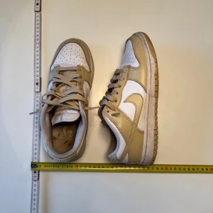 Nike Dunk Low beige och vita sneakers - Nike Dunk Low sneakers i beige och vitt med klassisk swoosh på sidorna. Skorna har snörning, platt sula och rund tå. Ovandelen är i skinn och textil, vilket ger en snygg kontrast mellan färgerna. Perfekta för dig som gillar stilrena och trendiga sneakers.