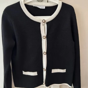 Ny Svart kofta med pärlknappar från Zara - Snygg svart kofta från Zara med kontrasterande vita detaljer vid krage, knappslå och fickor. Koftan har dekorativa pärlknappar och är långärmad. Materialet är stickad och ger en mjuk och skön känsla. Perfekt för dig som gillar klassisk stil med en twist.