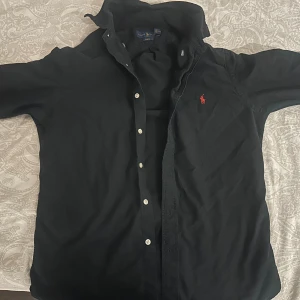 Svart skjorta från Ralph Lauren - Svart skjorta från Ralph Lauren i slim fit med knappar framtill. Storlek M pris kan diskuteras.