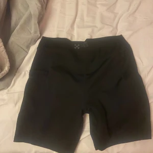 Svarta träningstights-shorts X-TRAIL - Svarta, tighta träningstights-shorts från X-TRAIL med midwaist-passform. De är gjorda i ett stretchigt syntetmaterial och har en diskret ficka på sidan. Perfekta för gymmet eller löprundan.